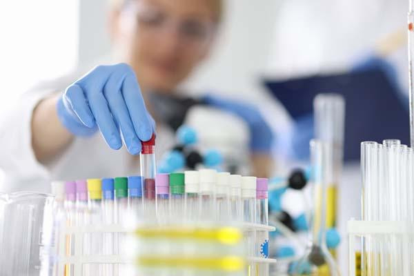 A rack of test tubes with different colored caps, with a gloved hand inserting a tube into the rack; in the background, out of focus, the lab tech's face is slightly visible